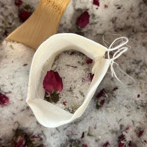May include: An off-white sachet filled with bath salts and a red rose bud. The sachet is tied with white string and rests on a bed of bath salts, dried herbs, and rose petals. A wooden scoop is in the background.