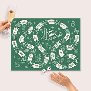 Puede incluir: Un juego de mesa verde con texto e ilustraciones en blanco, que incluye una pancarta que dice "Camp Emily Bachelorette Weekend". Una mano sostiene una copa de champán y hay dados en el tablero.