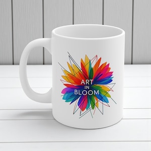 以下が含まれることがあります： 「ART IN BLOOM」の文字とカラフルな花のイラストが描かれた白いセラミックマグカップ。赤、オレンジ、黄色、緑、青、紫の色合いが使われています。マグカップはカーブしたハンドルと滑らかで光沢のある仕上がりです。