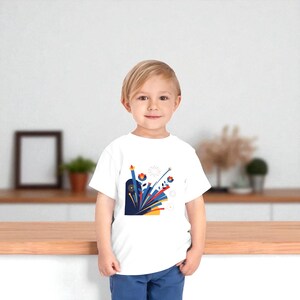以下が含まれることがあります： カラフルなグラフィックデザインが施された白いTシャツ。デザインには、赤、青、黄、白の様式化された花、線、形が含まれています。Tシャツは幼い子供が着用しています。