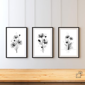 以下が含まれることがあります： 3つの額入りフローラルアートプリント。各黒いフレームのプリントは、幾何学的な形をした白黒のフローラルデザインが特徴です。プリントは白い壁を背景に木製の表面に表示されています。画像の右下隅には「ART IN BLOOM」というテキストがあります。