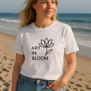 以下が含まれることがあります： 「ART IN BLOOM」の文字と黒い花のイラストがプリントされた白いTシャツ。モデルは青いジーンズを着用し、海を背景にしたビーチに立っています。