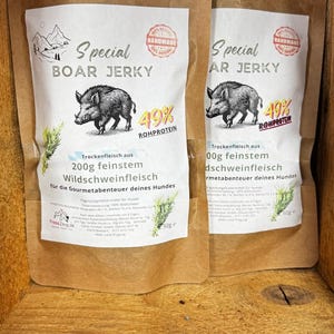 Könnte beinhalten: Zwei braune Papiertüten mit Boar Jerky für Hunde. Jede Tüte zeigt eine Zeichnung eines Wildschweins, den Text "Special Boar Jerky" und "49% Rohprotein". Die Tüten sind mit "200g feinstem Wildschweinfleisch" beschriftet.