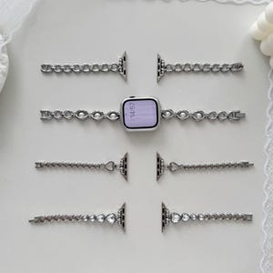 May include: Silver-toned watch bands with various decorative designs, including chain links, pearls, and heart-shaped accents. The watch face displays the time as 14:57. The bands are arranged on a white surface.