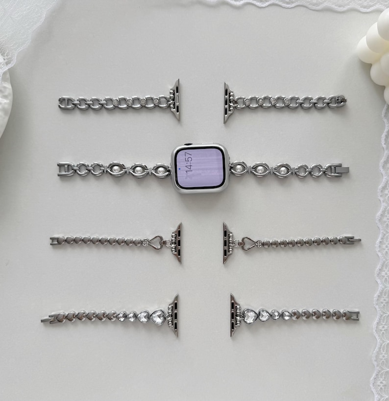May include: Silver-toned watch bands with various decorative designs, including chain links, pearls, and heart-shaped accents. The watch face displays the time as 14:57. The bands are arranged on a white surface.
