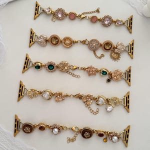May include: Gold-toned watch bands adorned with various charms, including flowers, hearts, and gemstones. The bands feature a mix of pink, green, and clear stones, with delicate chain accents. The bands are designed to fit a smartwatch.
