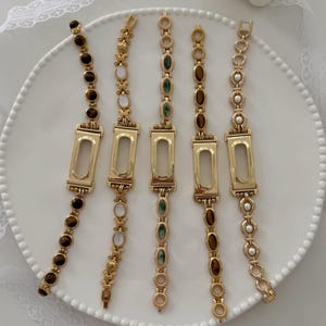 May include: Five gold-toned metal watch bands arranged vertically. Each band features a central rectangular frame and decorative links set with various stones, including brown tiger eye, white iridescent cabochons, green marbled stones, and white pearls.