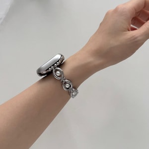 May include: A silver-toned smartwatch with a decorative band featuring a series of linked ovals and faux pearls. The watch is worn on a wrist, showcasing the design and style of the wearable tech accessory.