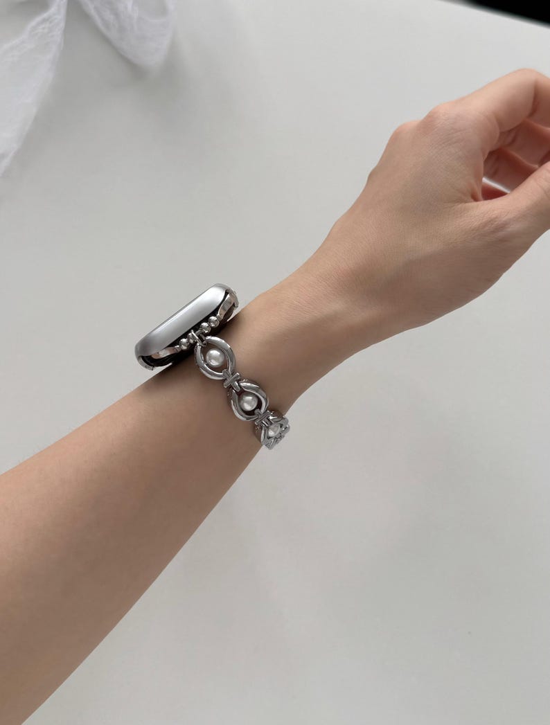 May include: A silver-toned smartwatch with a decorative band featuring a series of linked ovals and faux pearls. The watch is worn on a wrist, showcasing the design and style of the wearable tech accessory.