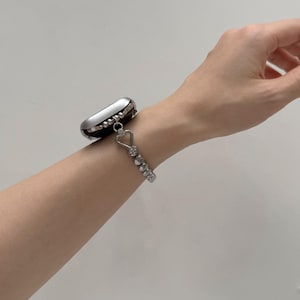 May include: A silver smartwatch with a beaded bracelet featuring a heart-shaped charm. The watch case is silver with a black border. The bracelet is made of small, round beads and connects to the watch with a heart-shaped link.