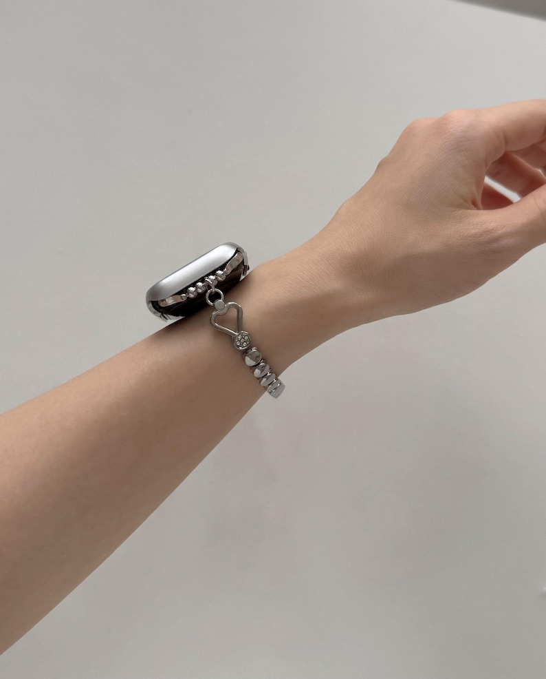 May include: A silver smartwatch with a beaded bracelet featuring a heart-shaped charm. The watch case is silver with a black border. The bracelet is made of small, round beads and connects to the watch with a heart-shaped link.