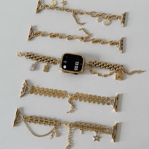 Könnte beinhalten: Fünf goldfarbene Apple Watch-Armbänder mit verschiedenen Ketten- und Charm-Designs. Ein Armband ist an einer goldenen Uhr mit einem schwarzen Bildschirm befestigt, der die Uhrzeit 13:55 anzeigt. Die Armbänder haben Charms wie Sterne, Monde und Münzen.