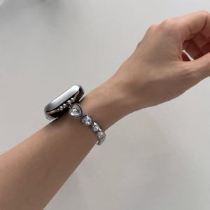 May include: A silver smartwatch with a decorative band featuring heart-shaped crystals. The band is attached to the watch and is worn on a wrist. The watch and band are made of a shiny, metallic material.