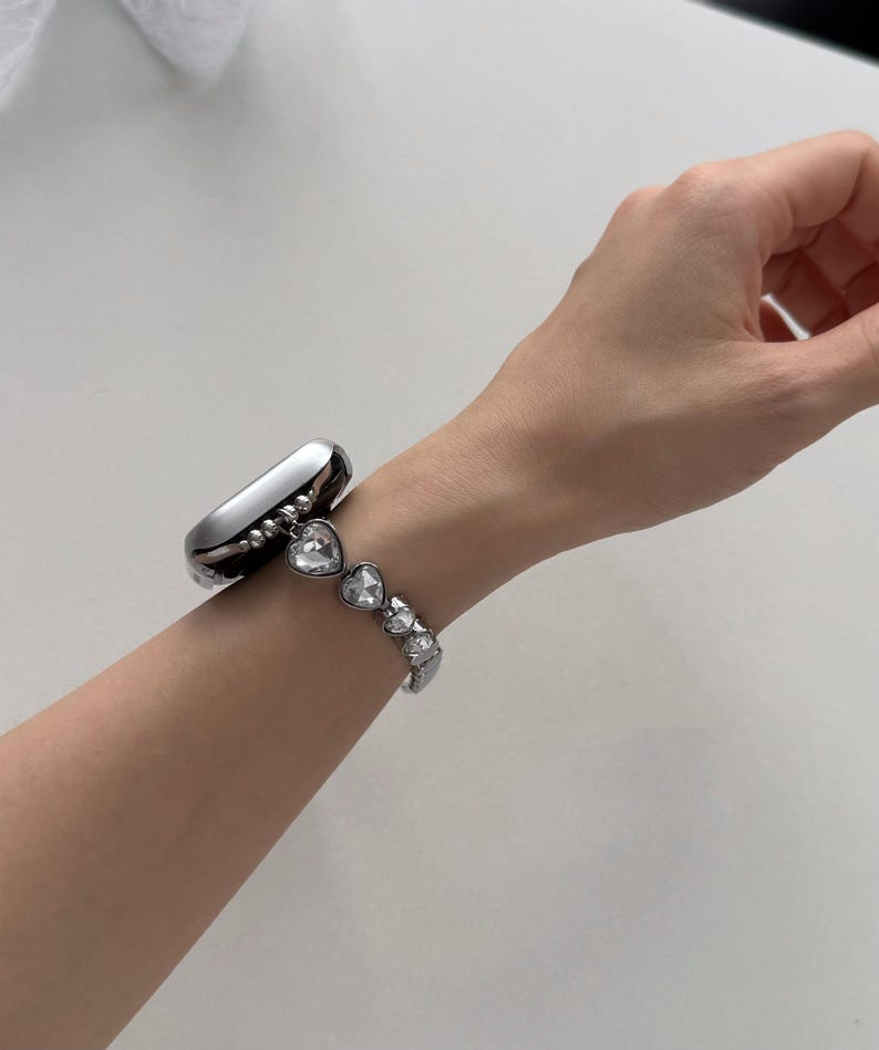 May include: A silver smartwatch with a decorative band featuring heart-shaped crystals. The band is attached to the watch and is worn on a wrist. The watch and band are made of a shiny, metallic material.