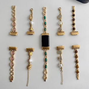 May include: Gold-toned watch band accessories with various decorative elements. The bands feature pink, green, and brown stones, along with star and heart-shaped charms. The accessories are displayed on a white surface.