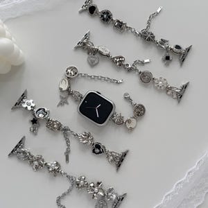 Peut inclure: Bracelets de montre argentés avec des breloques décoratives, notamment des fleurs, des cœurs et des étoiles. Les bracelets sont présentés avec une montre intelligente argentée à écran noir. Les breloques ont des accents noirs et blancs.