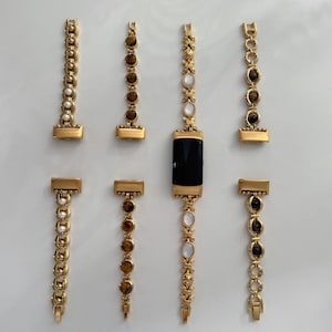 May include: A collection of gold-toned metal watch bands for a fitness tracker, featuring various decorative links including white pearls, brown tiger eye stones, and iridescent oval cabochons. One band is attached to a black rectangular tracker.