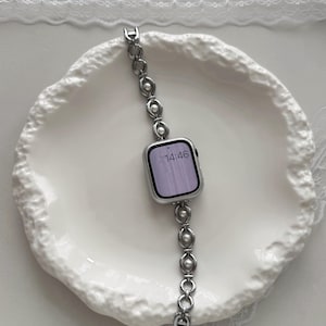 May include: A silver smartwatch with a linked band featuring pearl accents. The watch face displays the time as 14:46. The watch is set on a white, textured plate.