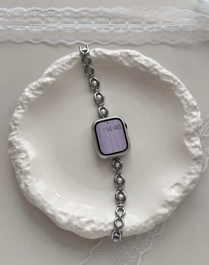 May include: A silver smartwatch with a linked band featuring pearl accents. The watch face displays the time as 14:46. The watch is set on a white, textured plate.