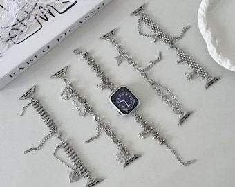 May include: Silver-toned watch bands with various chain and charm designs. One band is attached to a square-faced watch with a dark blue watch face. The bands feature decorative elements like stars, beads, and heart-shaped charms. The background includes a book with the text "PABLO PICASSO".