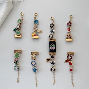 Könnte beinhalten: Goldfarbene Armbanduhren-Anhänger mit farbenfrohen dekorativen Elementen. Die Anhänger weisen verschiedene Designs auf, darunter Blumenmuster, Edelsteine und aufwendige Details. Einige Anhänger haben baumelnde Ketten.