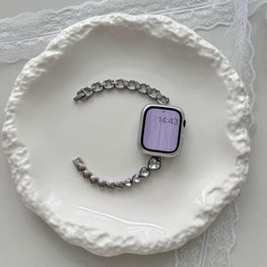 May include: Silver-toned Apple Watch with a decorative band featuring a chain and heart design. The watch face displays the time 14:43. The watch and band are displayed on a white, textured plate.