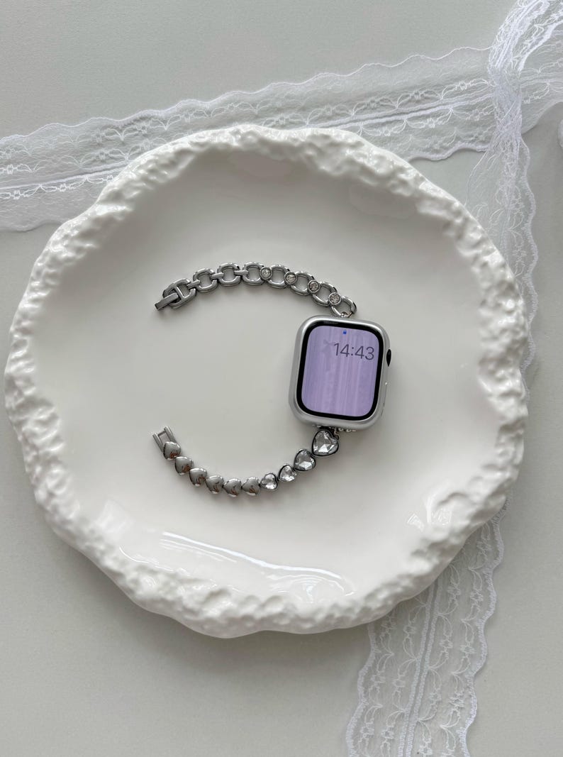 May include: Silver-toned Apple Watch with a decorative band featuring a chain and heart design. The watch face displays the time 14:43. The watch and band are displayed on a white, textured plate.