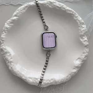 May include: A silver smartwatch with a heart-shaped link band rests on a white, textured plate. The watch face displays the time 14:47. The band is composed of small, heart-shaped links and round links.
