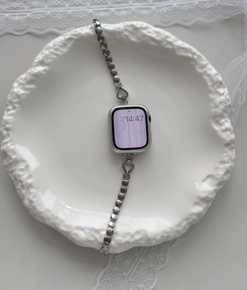 May include: A silver smartwatch with a heart-shaped link band rests on a white, textured plate. The watch face displays the time 14:47. The band is composed of small, heart-shaped links and round links.