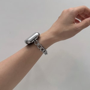 May include: A silver smartwatch with a decorative chain-link band is worn on a wrist. The band features a series of interconnected, open-circle links. The watch case is a rounded rectangle. The background is a plain, light color.