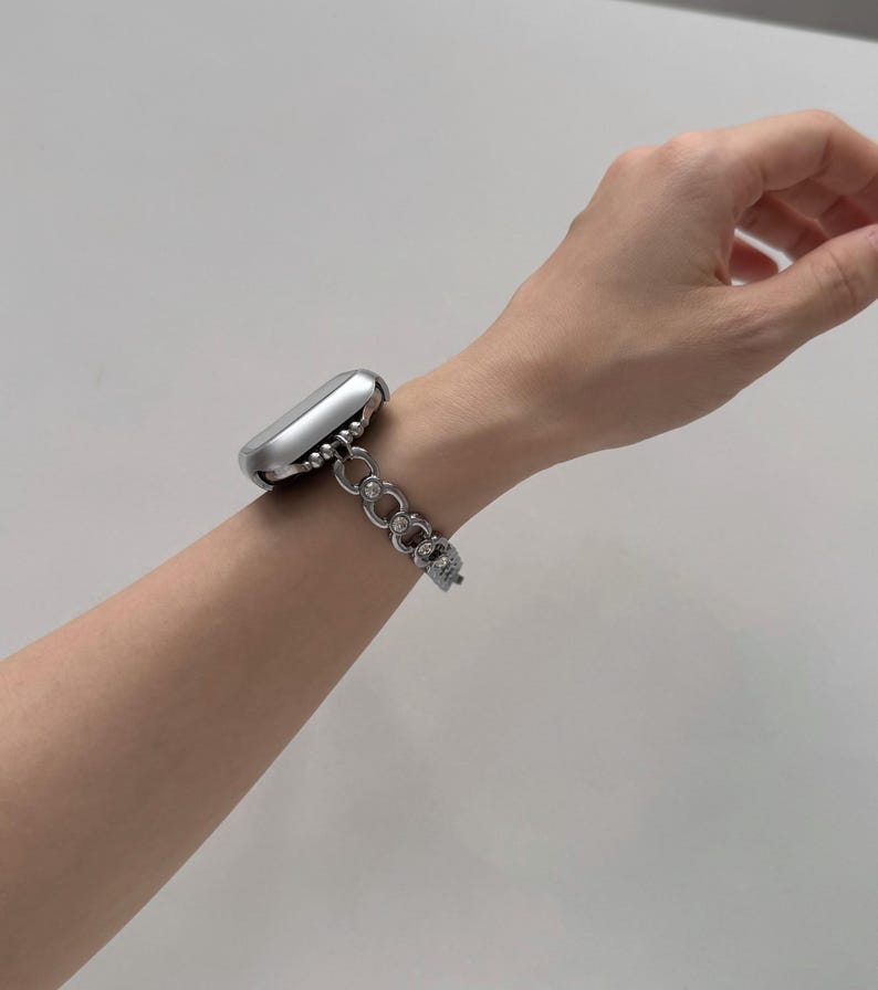May include: A silver smartwatch with a decorative chain-link band is worn on a wrist. The band features a series of interconnected, open-circle links. The watch case is a rounded rectangle. The background is a plain, light color.