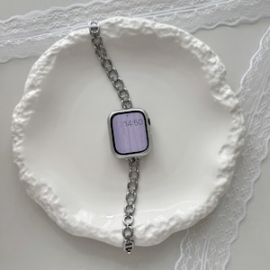 May include: A silver smartwatch with a linked band, resting on a white, textured plate. The watch face displays the time 14:50. The band is made of small, connected links. The plate has a wavy, organic shape, and is surrounded by white lace.