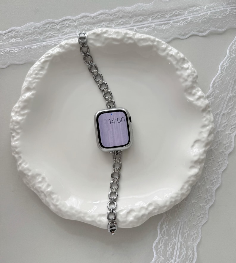 May include: A silver smartwatch with a linked band, resting on a white, textured plate. The watch face displays the time 14:50. The band is made of small, connected links. The plate has a wavy, organic shape, and is surrounded by white lace.