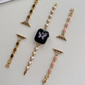 May include: An Apple Watch with a black screen showing a butterfly design, surrounded by gold-coloured watch straps. The straps feature decorative elements such as pearls, pink stones, and brown stones. The watch displays the time 15:07.