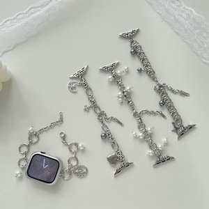 May include: Silver Apple Watch straps with decorative charms and faux pearls. The straps feature various charms, including hearts, stars, and small beads. One strap is attached to a silver Apple Watch.