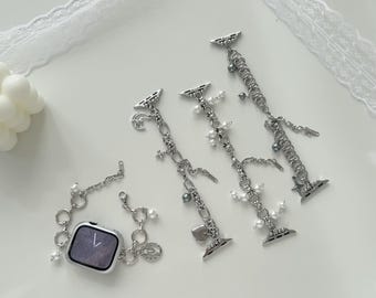 May include: Silver-toned Apple Watch bands with decorative charms and faux pearls. The bands feature various charms, including hearts, stars, and small beads. One band is attached to a silver Apple Watch.