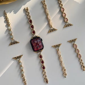 Puede incluir: Correas doradas para Apple Watch con detalles de piedras preciosas. Las correas presentan piedras rojas, rosas y crema, y se muestran con un Apple Watch negro que muestra la hora 15:56. Las correas son intercambiables.