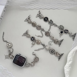 Peut inclure: Bracelets de montre argentés avec des breloques décoratives, notamment des étoiles, des perles et des motifs floraux. L'un des bracelets est fixé à une montre connectée argentée affichant l'heure. Les bracelets présentent diverses formes et textures.
