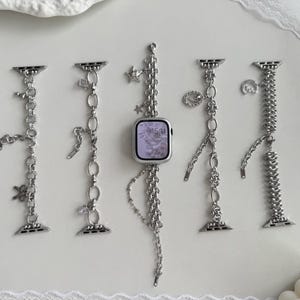 Peut inclure: Bracelets de montre argentés avec divers modèles de chaînes et de breloques. Les bracelets présentent des éléments décoratifs tels que des étoiles, des fleurs et des pendentifs ovales. Un bracelet est attaché à une montre connectée argentée affichant l'heure.