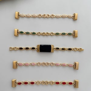 Könnte beinhalten: Fünf goldfarbene Armbänder mit verschiedenen farbigen, oval geformten Steinen. Die Steine umfassen Creme, Grün, Schwarz, Rosa und Rot. Jedes Armband hat an jedem Ende einen rechteckigen Verschluss.