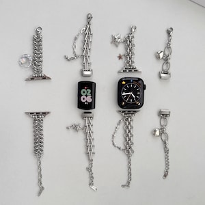 Op de afbeelding: Zilverkleurige horlogebandjes en bedels worden gepresenteerd op een witte ondergrond. De bandjes hebben ketting- en bedelontwerpen, met ook een smartwatch en een fitnesstracker. De wijzerplaten van de horloges tonen de tijd en datum.