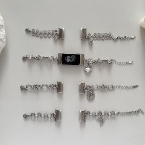 Peut inclure: Bracelets de montre argentés avec divers motifs de chaînes décoratives, dont des cœurs, des croix et des perles. L'un des bracelets présente un écran numérique noir. Les bracelets sont posés sur une surface blanche.
