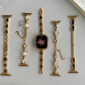 May include: Gold-toned watch bands with decorative charms and a square-faced watch. The bands feature various designs, including chain links, gemstones, and heart-shaped accents. The watch face displays a red and black design.