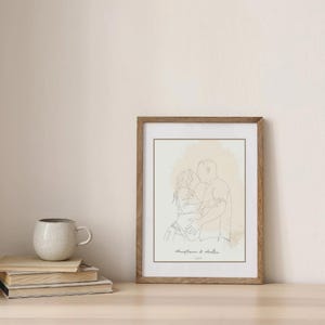 以下が含まれることがあります： 抱きしめ合ってキスをしているカップルの線画プリントが額装されています。アートワークはミニマリストスタイルで、ニュートラルなカラーパレットです。フレームは木製です。プリントは、本とマグカップが置かれたテーブルの上にあります。