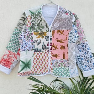 Puede incluir: Chaqueta patchwork con diseño de cuello en V, con una variedad de patrones florales y geométricos en tonos verdes, rosas y azules. La chaqueta tiene un ribete blanco y está colgada de una percha plateada.