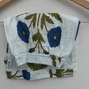 Pode incluir: Calças de pijama brancas com um padrão floral azul e folhas verdes, penduradas num cabide de madeira. As calças têm uma cintura elástica e são feitas de um tecido leve. O design floral apresenta grandes flores azuis.