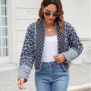May include: A blue and white floral quilted jacket with a collarless design and long sleeves. The jacket has striped cuffs. The woman is wearing a white top and blue jeans. She is also wearing sunglasses and a necklace.