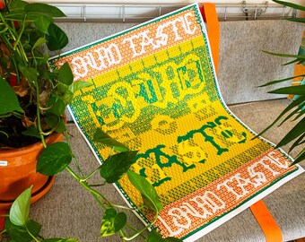May include: A colorful textile art piece with the words "SOUND TASTE" in white and green, set against a yellow and green background. The artwork is displayed on a gray couch with orange straps, surrounded by green plants.