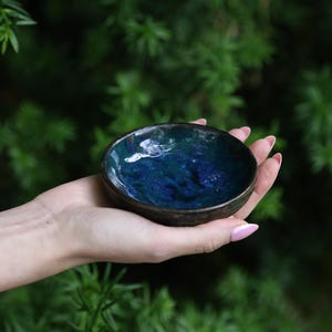 May include: A small, handmade ceramic bowl with a dark blue and teal interior. The bowl has a dark brown rim and is held in a hand against a blurred green background. The bowl's interior has a glossy, marbled appearance.