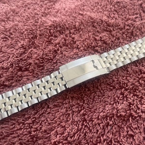 Può includere: Un cinturino per orologio in metallo color argento caratterizzato da un design a maglie multiple con una chiusura centrale lucida. Il cinturino è composto da file alternate di maglie spazzolate e lucide, creando un aspetto strutturato.